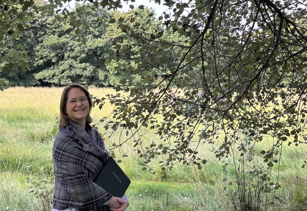 Fleur standing in a meadow with a lilac tartan shawl and ceremony folder under the boughs of a birch tree
