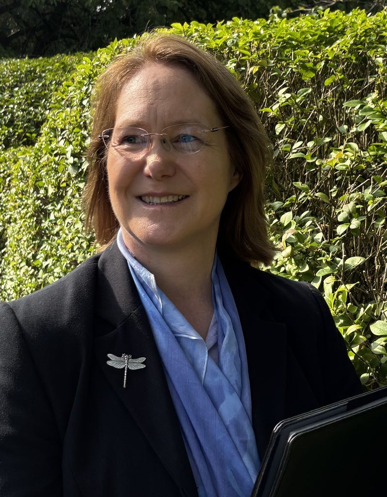 Fleur smiling and holding a folder, wearing a smart suit with a sky-blue scarf and silver dragonfly brooch. 